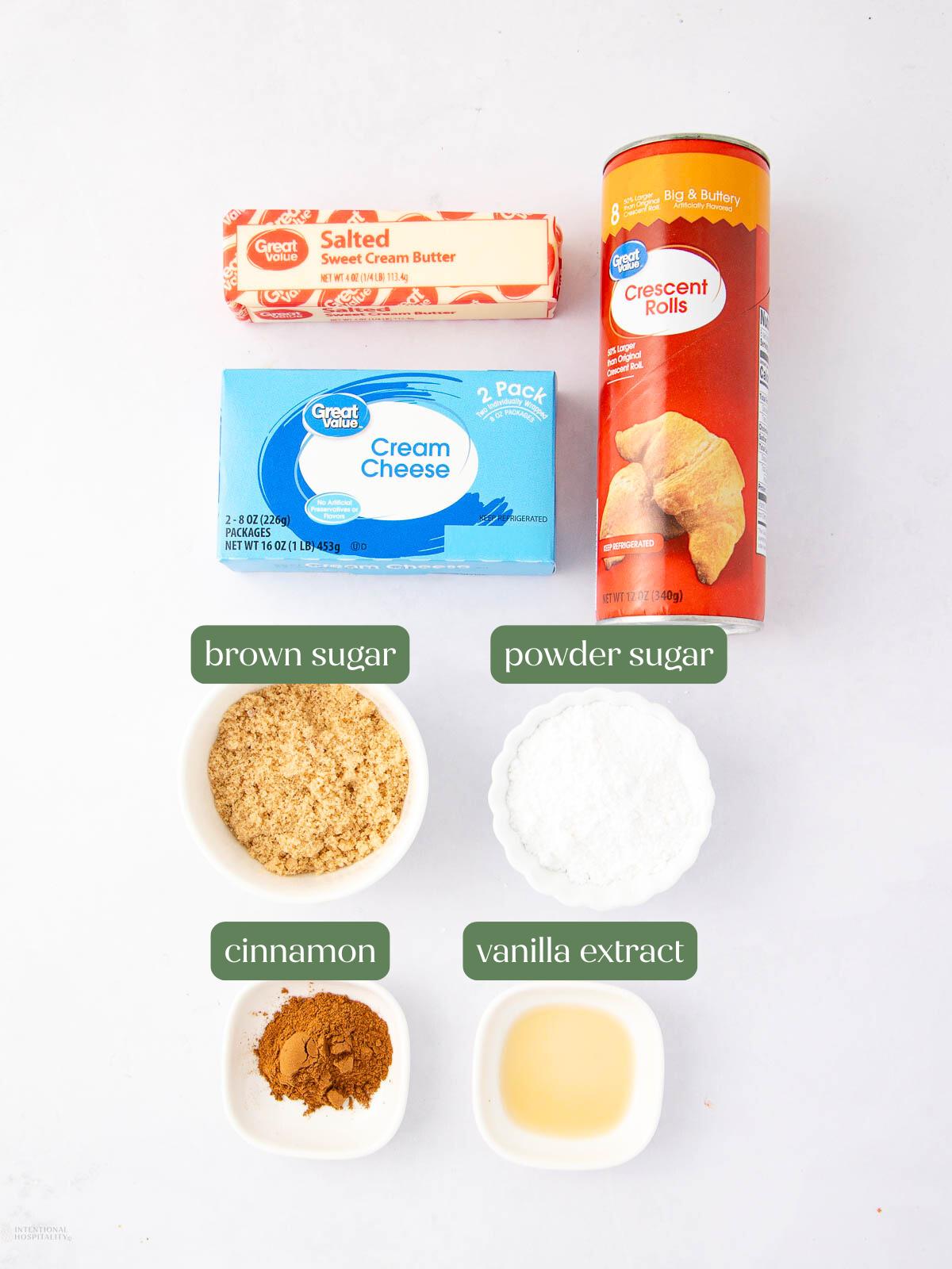 Ingredients for a recipe are arranged on a white surface: salted butter, cream cheese, crescent rolls, brown sugar, powdered sugar, cinnamon, and vanilla extract in small bowls, each labeled.