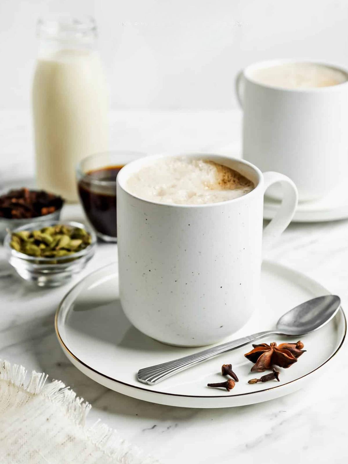 A white mug of frothy chai latte on a plate with a spoon, star anise, and cloves, next to bowls of spices, a glass of milk, and another mug in the background on a marble surface.