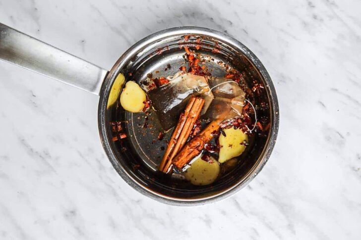 A saucepan on a marble surface containing water, cinnamon sticks, ginger slices, tea bags, and red pepper flakes.
