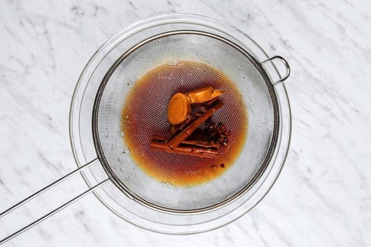 A metal strainer over a glass bowl holds cinnamon sticks, ginger, and spices, with dark liquid strained below on a white marble surface.