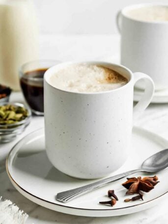 A white mug filled with frothy chai latte on a white saucer with a spoon, star anise, and cloves. In the background are spices in small bowls, a cup of coffee, and another mug on a white surface.