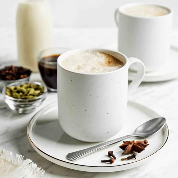 A white mug filled with frothy chai latte on a white saucer with a spoon, star anise, and cloves. In the background are spices in small bowls, a cup of coffee, and another mug on a white surface.