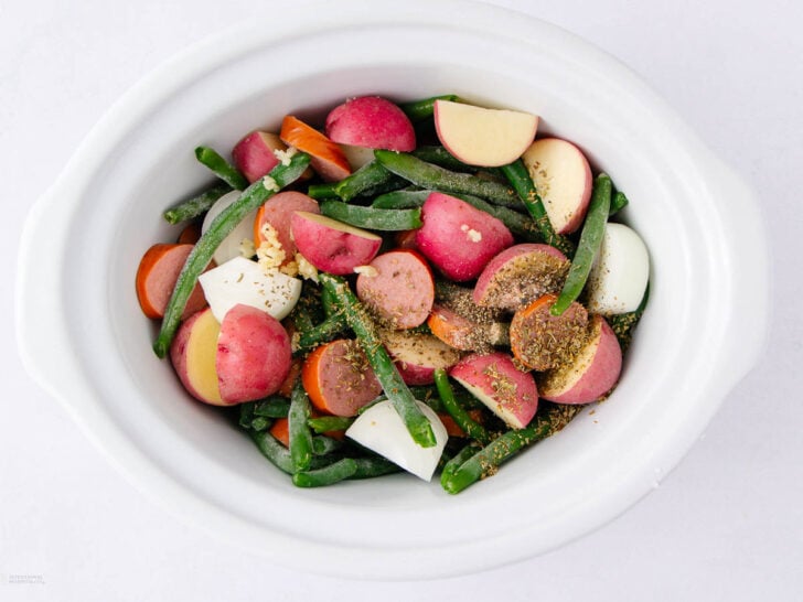 A white slow cooker filled with chopped red potatoes, carrots, green beans, and onions, sprinkled with herbs and spices, ready to be cooked.