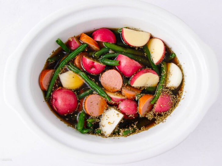 A white slow cooker filled with sliced red potatoes, green beans, carrot pieces, and herbs soaking in a broth.