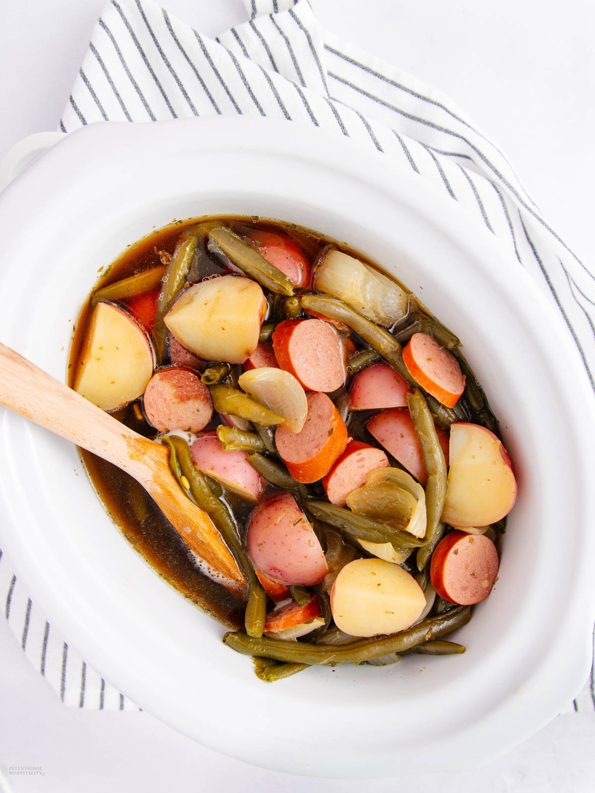A white oval dish filled with sliced sausage, potatoes, carrots, green beans, and onions in broth sits on a striped cloth. A wooden spoon rests inside the dish.