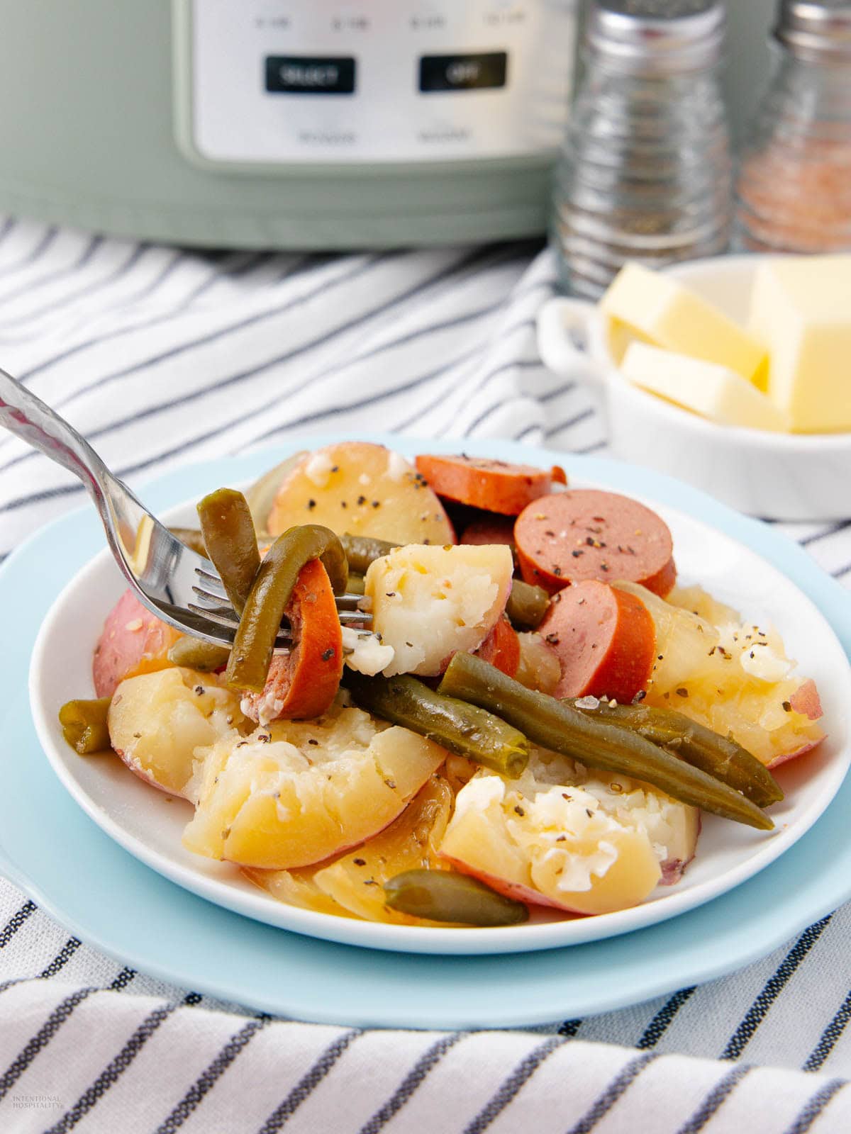 A plate of sliced sausage, potatoes, green beans, and onions, with a fork picking up a bite. There’s a striped cloth underneath, a dish of butter, salt and pepper shakers, and a slow cooker in the background.