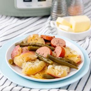 A plate of sliced potatoes, green beans, and sausage, seasoned with pepper, sits on a white dish. In the background are butter cubes, salt and pepper shakers, and a slow cooker, all on a striped cloth.