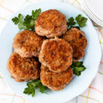 A plate of six browned sausage patties is arranged on a light blue dish, garnished with fresh parsley leaves, and set on a light-colored, checkered tablecloth near a stack of white plates.