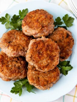 A plate of six browned sausage patties is arranged on a light blue dish, garnished with fresh parsley leaves, and set on a light-colored, checkered tablecloth near a stack of white plates.