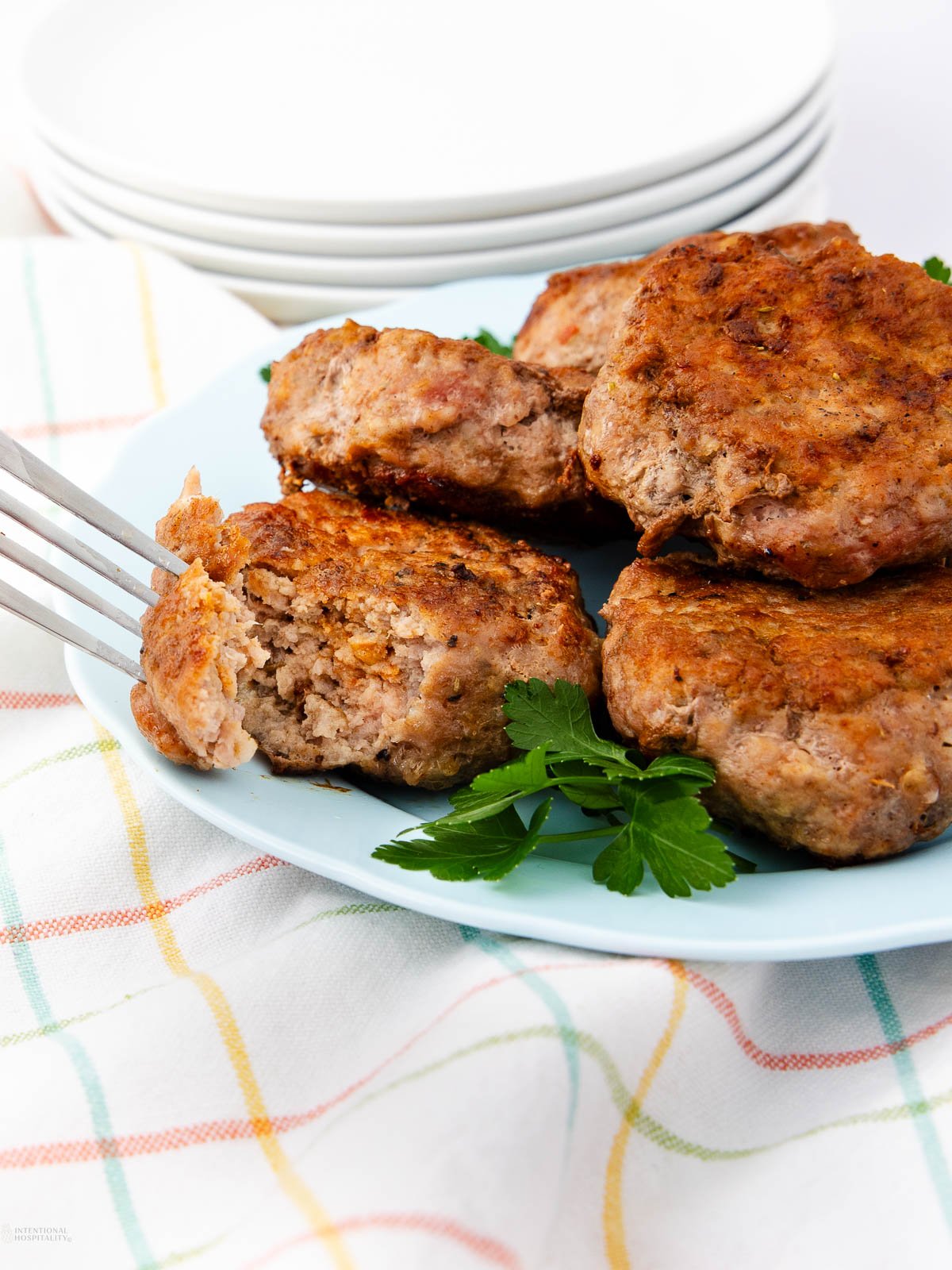Four browned meat patties on a light blue plate garnished with fresh parsley, with a fork piercing one patty. A stack of white plates and a checkered cloth are visible in the background.