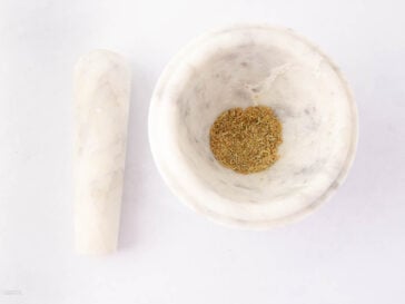 A marble mortar and pestle on a white surface. The mortar contains a small amount of ground spices or herbs, while the pestle rests beside it.