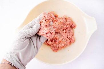 A hand in a white glove holds a portion of raw ground meat above a white mixing bowl containing more of the meat, against a white background.