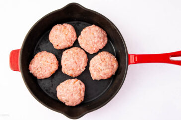 A red-handled skillet containing seven raw, round sausage patties arranged in a single layer on a white background.