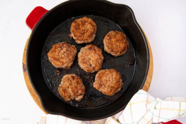 Six cooked sausage patties are arranged in a black cast iron skillet, which is placed on a wooden board. A white kitchen towel with a pastel plaid pattern is draped beside the skillet.