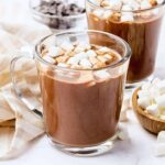 Two glass mugs filled with hot chocolate topped with mini marshmallows, placed on a white surface with a striped cloth, chocolate chips, and a small wooden bowl of marshmallows nearby.