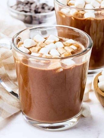 Two glass mugs filled with hot chocolate topped with mini marshmallows, placed on a white surface with a striped cloth, chocolate chips, and a small wooden bowl of marshmallows nearby.