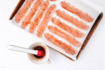 A baking tray lined with parchment paper holds several strips of uncooked vegan bacon, next to a cup with a red basting brush resting inside, filled with a dark marinade.
