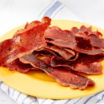 Crispy slices of vegan bacon arranged on a yellow plate, placed on a white cloth with blue stripes.
