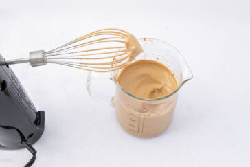 A metal whisk attached to a hand mixer is held above a glass measuring cup filled with whipped coffee on a white background. The coffee has a thick, creamy texture.