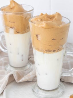 Two clear glass mugs filled with iced milk, topped with a thick, frothy coffee foam, sit on a light-colored cloth. The drinks are Dalgona coffee, with the coffee layer contrasting against the white milk.
