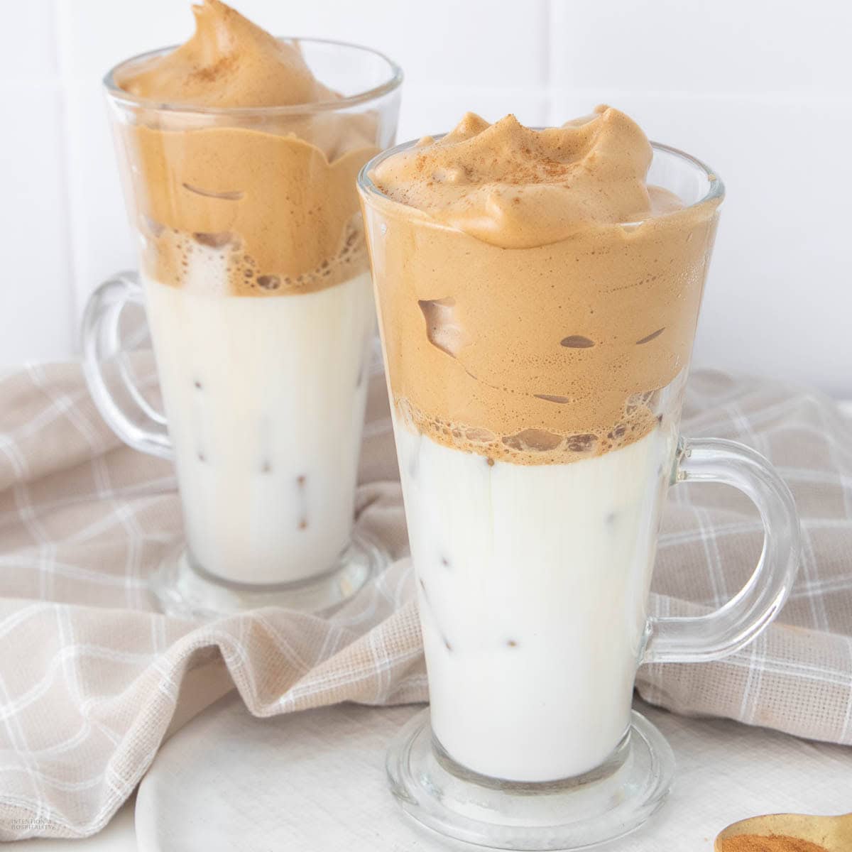 Two clear glass mugs filled with iced milk, topped with a thick, frothy coffee foam