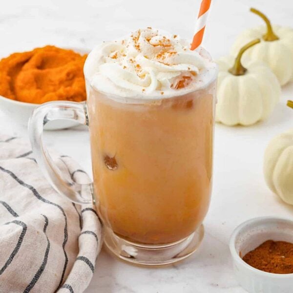 Iced Pumpkin latte topped with whipped cream and cinnamon in a clear glass mug with a striped straw, surrounded by small white pumpkins, a bowl of pumpkin puree, and a striped cloth.
