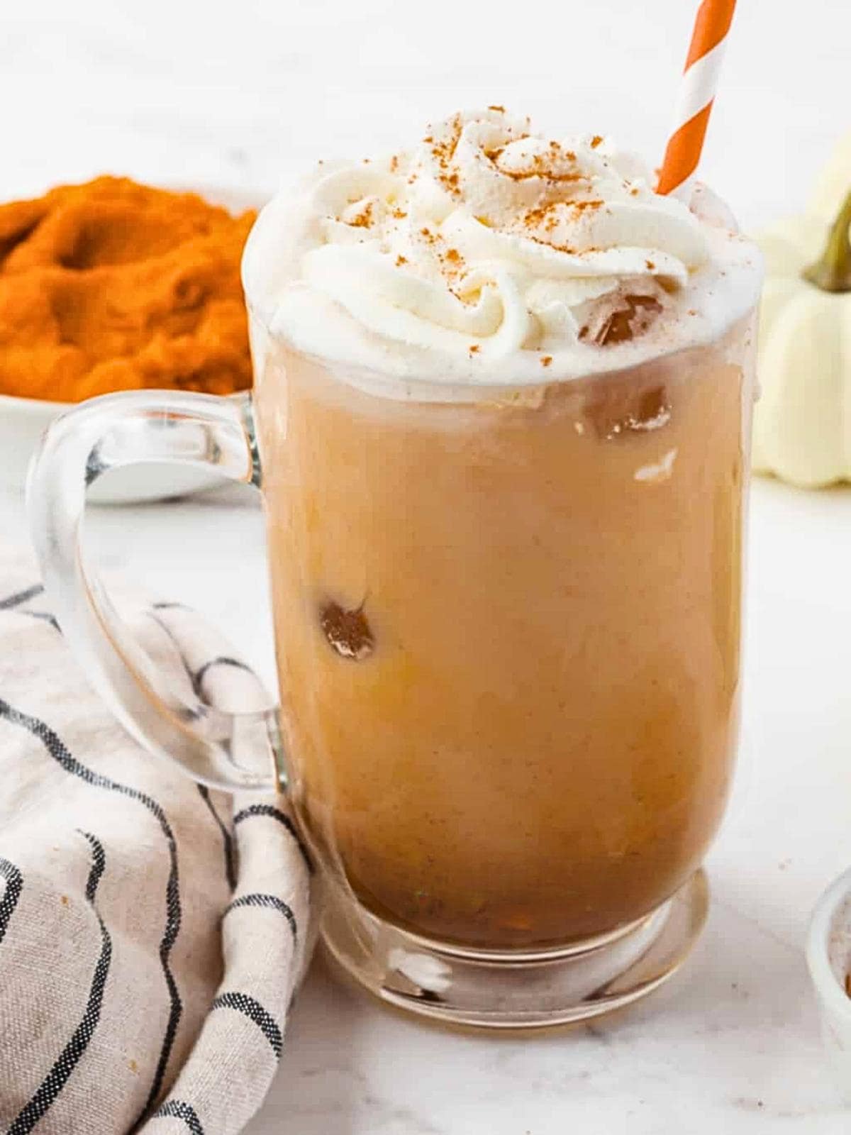 A glass mug of iced coffee topped with whipped cream and sprinkled with cinnamon, served with a striped straw. A bowl of pumpkin puree and a striped cloth are visible in the background.