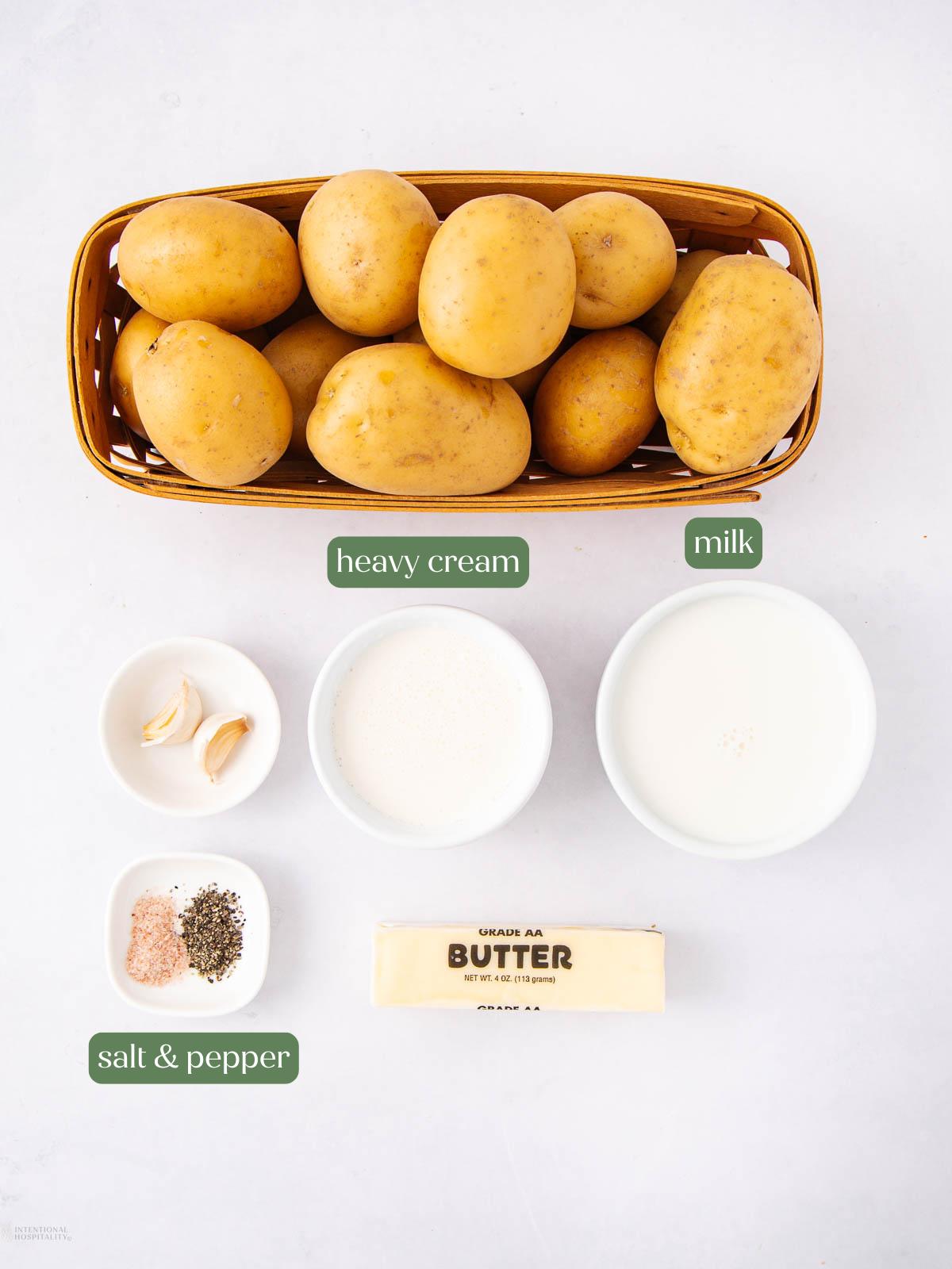 A basket of whole potatoes, a stick of butter, a bowl of heavy cream, a bowl of milk, a small bowl of garlic cloves, and a dish with salt and pepper are arranged on a white surface.