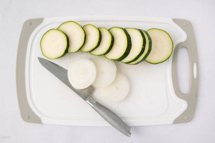 A cutting board with sliced zucchini and onion, a knife resting on the board. The zucchini slices are stacked, and the onion slices are arranged beside the knife.