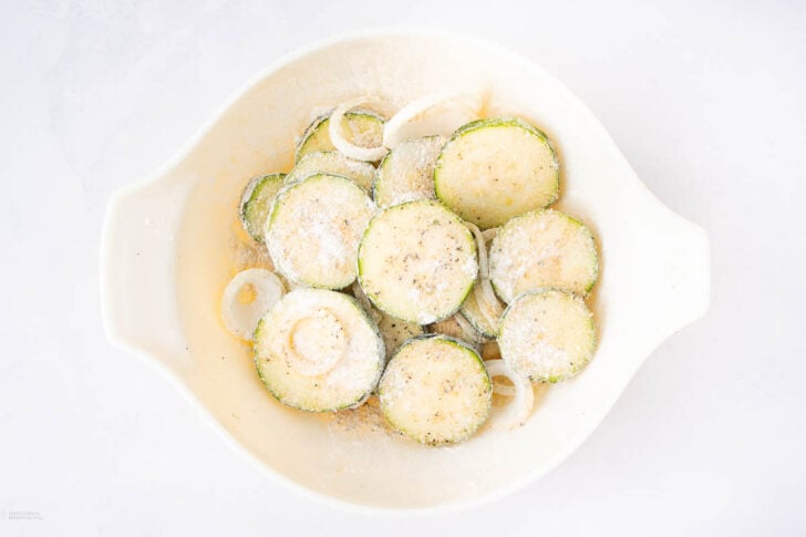 Sliced zucchini and onion rings in a white bowl, sprinkled with grated cheese and pepper, ready for cooking.
