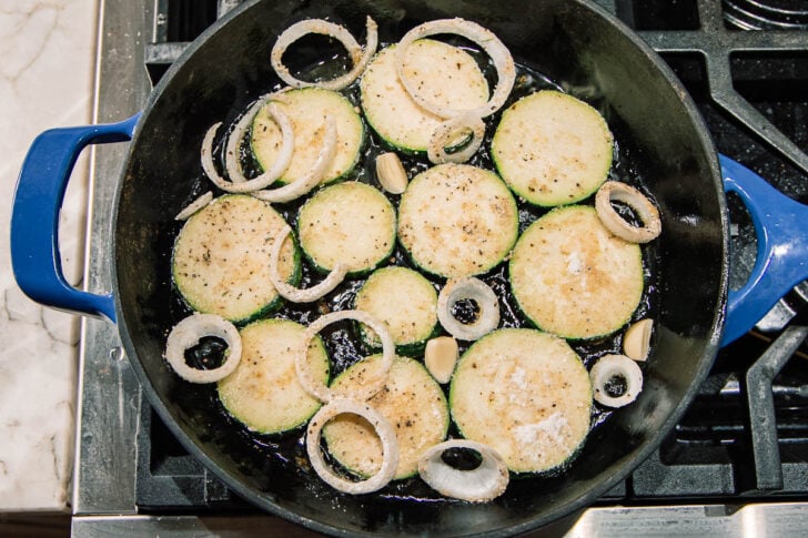 Sliced zucchini, onion rings, and garlic cloves are cooking in a blue cast iron skillet on a stovetop. The vegetables are seasoned and arranged in a single layer.