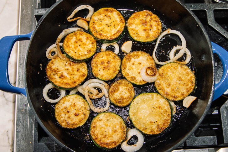 Sliced zucchini rounds, onion rings, and garlic cloves are being sautéed in a blue skillet on a stovetop. The zucchini slices are golden brown and appear crispy.
