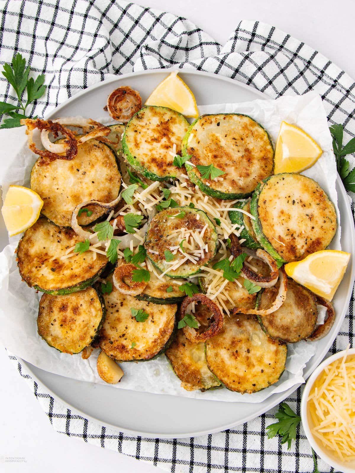 A plate of golden-brown fried zucchini slices garnished with fresh parsley, grated cheese, and crispy onions, served with lemon wedges on parchment paper. A checkered cloth and a bowl of cheese are nearby.