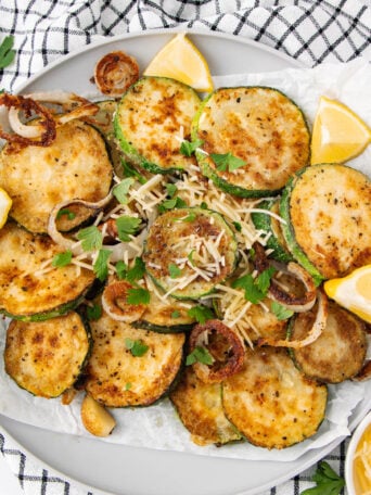 A plate of golden-brown, breaded fried zucchini slices garnished with parsley, grated cheese, and lemon wedges. Sautéed onions are scattered on top, and the plate sits on a black-and-white checkered cloth.