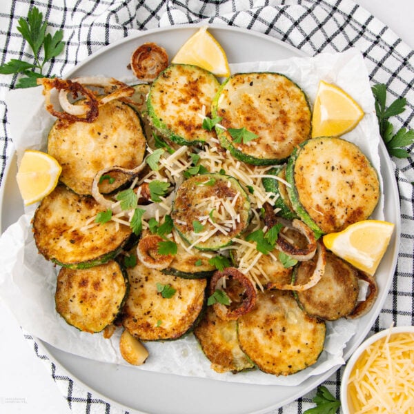 A plate of golden-brown, breaded fried zucchini slices garnished with parsley, grated cheese, and lemon wedges. Sautéed onions are scattered on top, and the plate sits on a black-and-white checkered cloth.