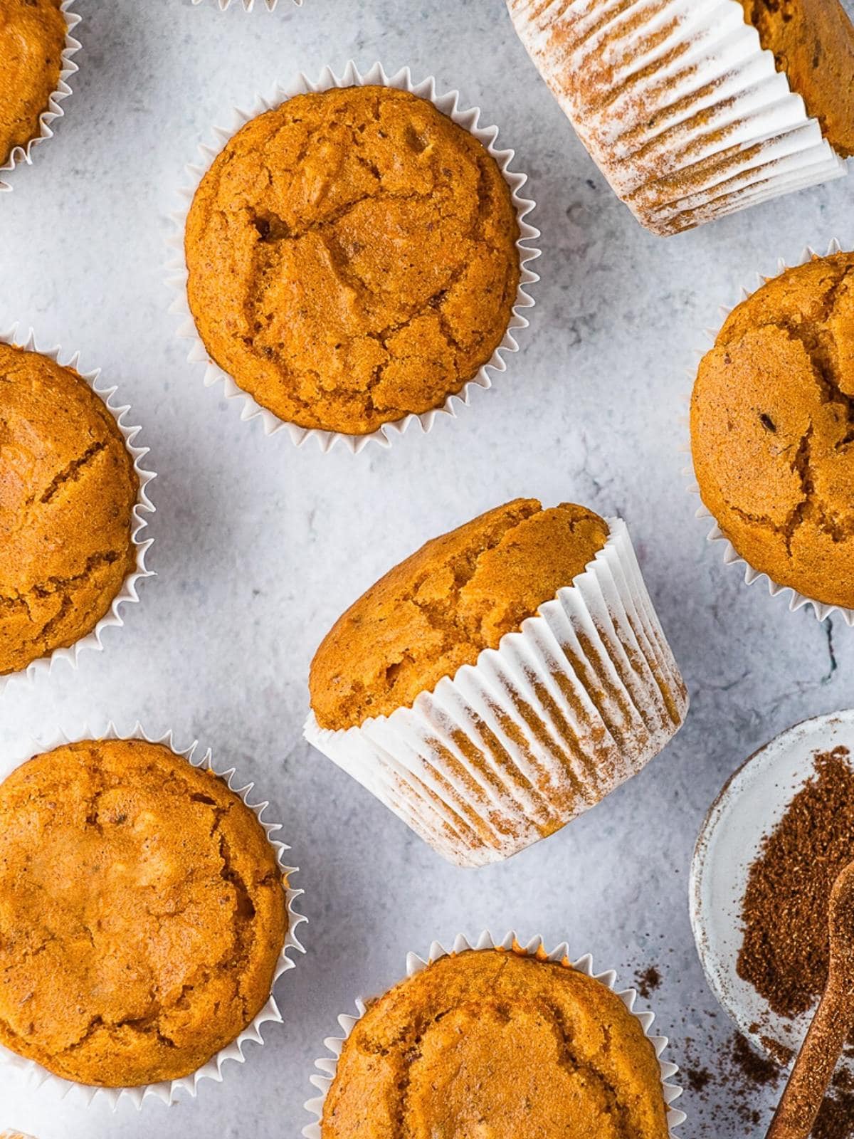 Pumpkin muffins in white paper liners are arranged on a light surface, with a bowl of ground spices and a dusting of spice nearby. The muffins have a golden-brown color and slightly cracked tops.