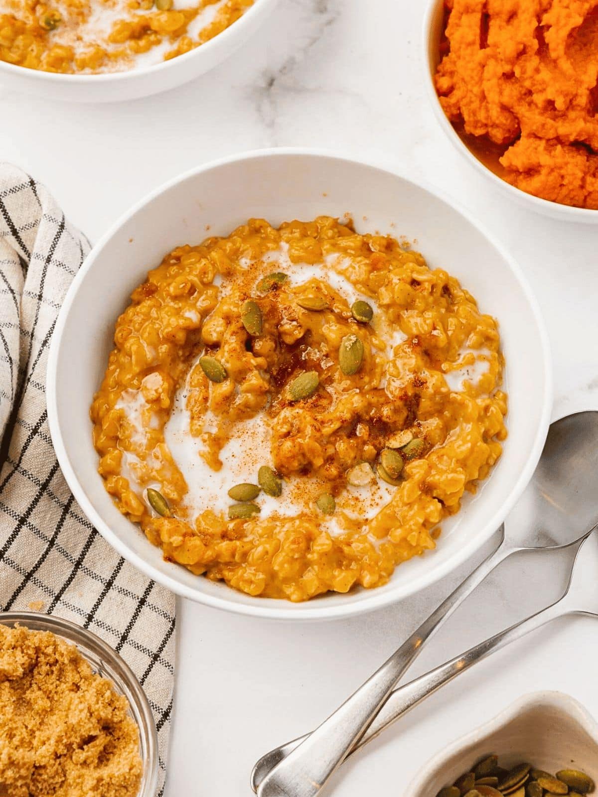A white bowl filled with pumpkin oatmeal, topped with a swirl of cream, pepitas, and a sprinkle of cinnamon. A spoon, striped napkin, brown sugar, and a bowl of mashed pumpkin are nearby.