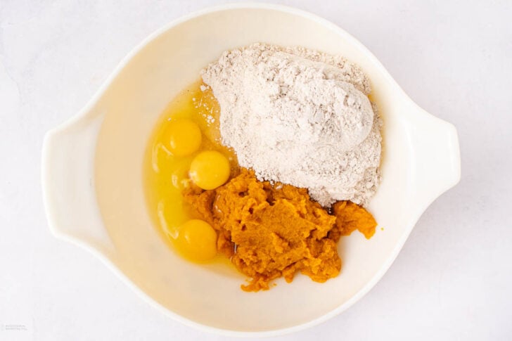 A white mixing bowl containing three cracked eggs, a mound of flour, and a portion of pumpkin puree, all unmixed, on a white surface.