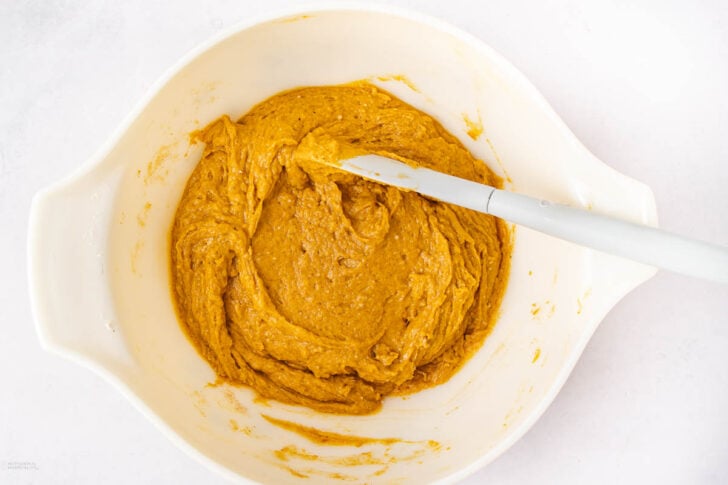 A white mixing bowl filled with thick pumpkin-colored batter, with a white spatula resting on the edge of the bowl. The bowl is placed on a white surface.