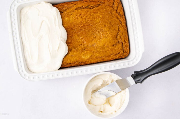 Rectangular cake in a white baking dish, partially frosted with white icing. A bowl of icing and a spatula with some icing on it are next to the dish on a white surface.