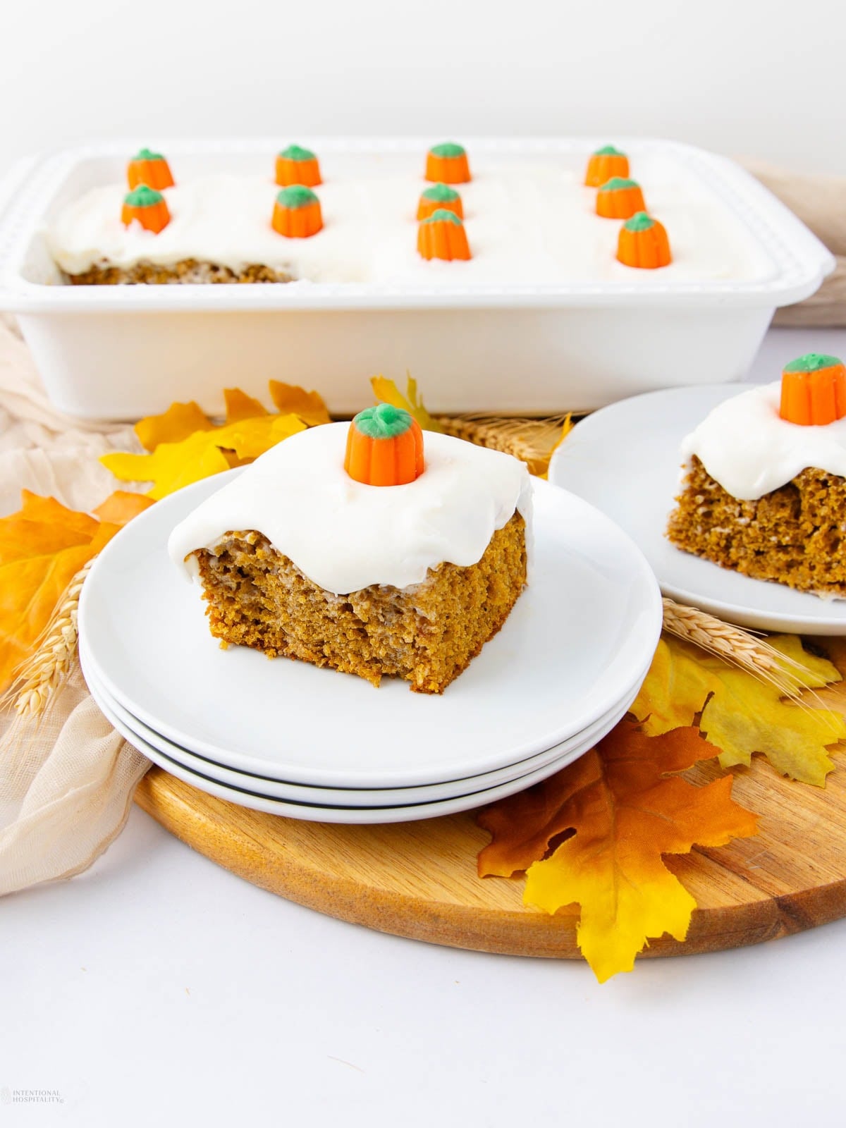 A slice of pumpkin cake with white frosting and a candy pumpkin on top sits on a plate. The rest of the cake, also frosted and topped with candy pumpkins, is in a baking dish behind it, surrounded by autumn leaves.