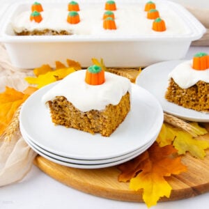 A slice of pumpkin cake with white frosting and a candy pumpkin on top sits on a white plate. The cake is surrounded by autumn leaves, and a baking dish with more cake is in the background.