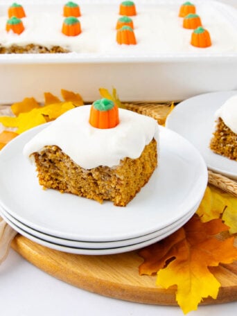 A slice of pumpkin cake with white frosting and a candy pumpkin on top sits on a white plate. The cake is surrounded by autumn leaves, and a baking dish with more cake is in the background.