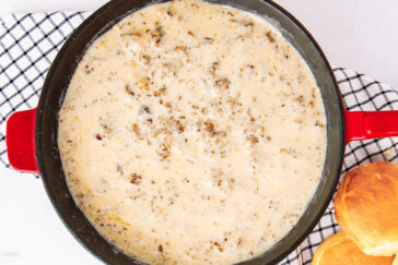 A red skillet filled with creamy sausage gravy sits on a black-and-white checkered cloth, with two biscuits placed nearby on the right side.