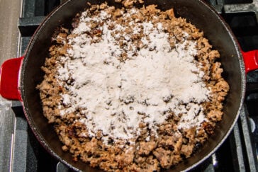 Ground meat cooking in a red skillet on a stove, topped with a layer of white flour sprinkled over the meat.