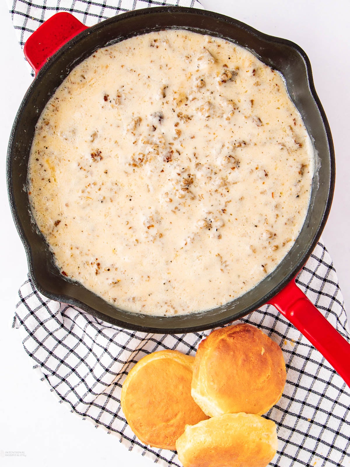 A skillet of creamy sausage gravy sits on a checkered cloth next to two golden brown biscuits.