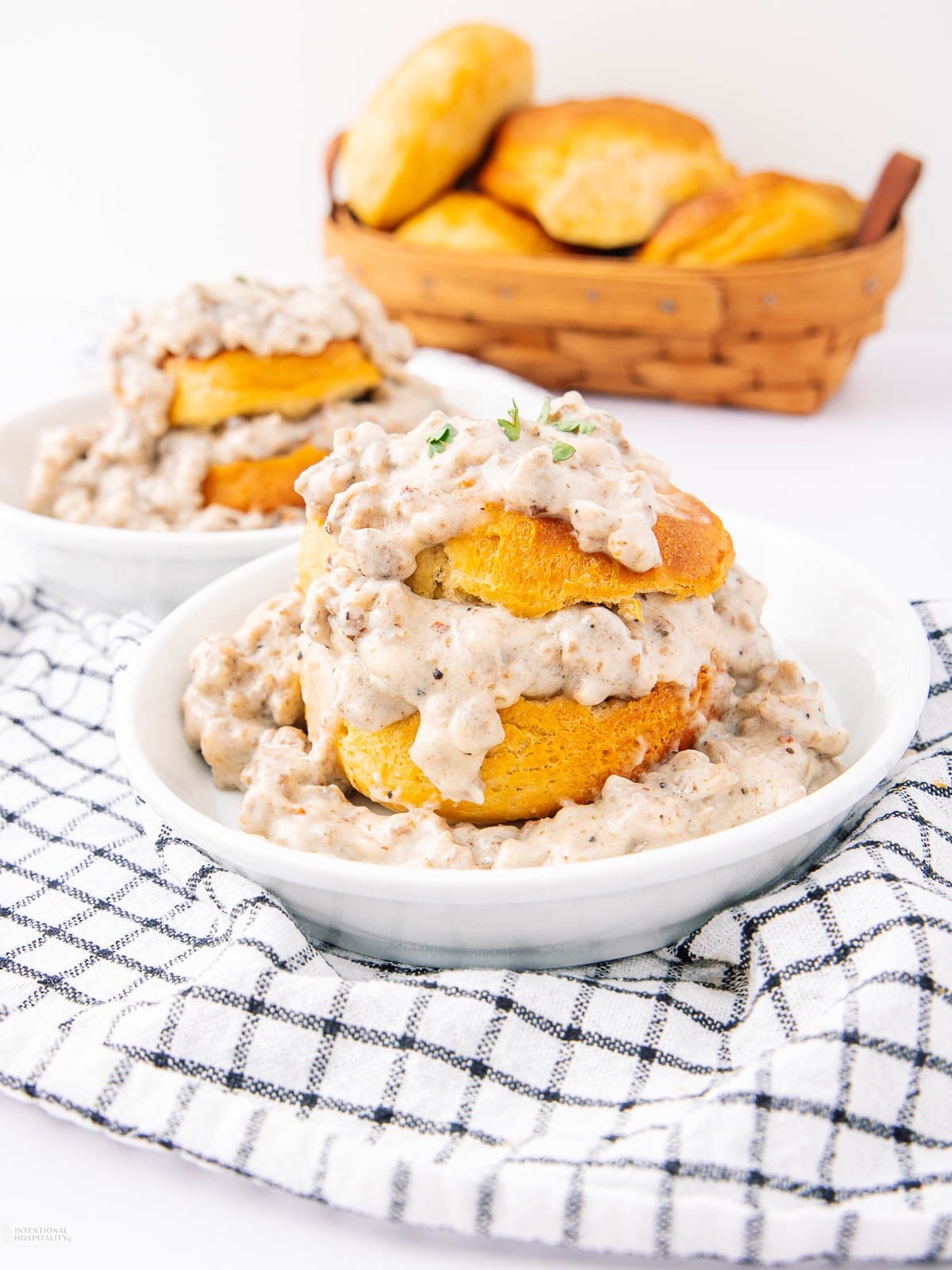 Two white bowls filled with golden biscuits topped with creamy sausage gravy, set on a checkered cloth. A basket of extra biscuits is in the background.