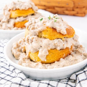 A bowl of biscuits cut in half and stacked, generously topped and surrounded by thick sausage gravy, sits on a white plate with a checkered cloth underneath. Another serving is visible in the background.