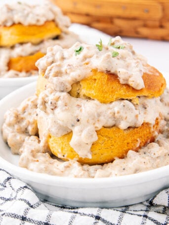 A bowl of biscuits cut in half and stacked, generously topped and surrounded by thick sausage gravy, sits on a white plate with a checkered cloth underneath. Another serving is visible in the background.