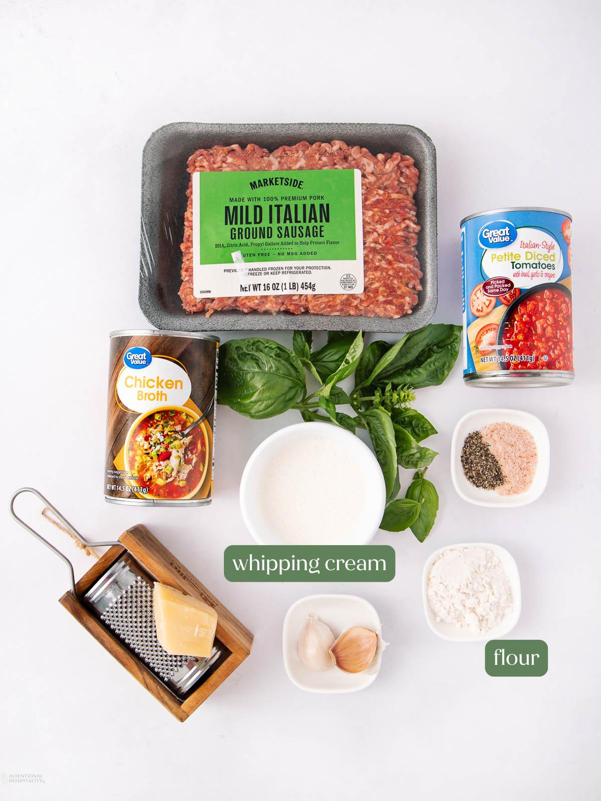 A flat lay of ingredients on a white surface: ground mild Italian sausage, canned diced tomatoes, chicken broth, whipping cream, fresh spinach, grated Parmesan, garlic cloves, flour, salt, pepper, and Italian seasoning.